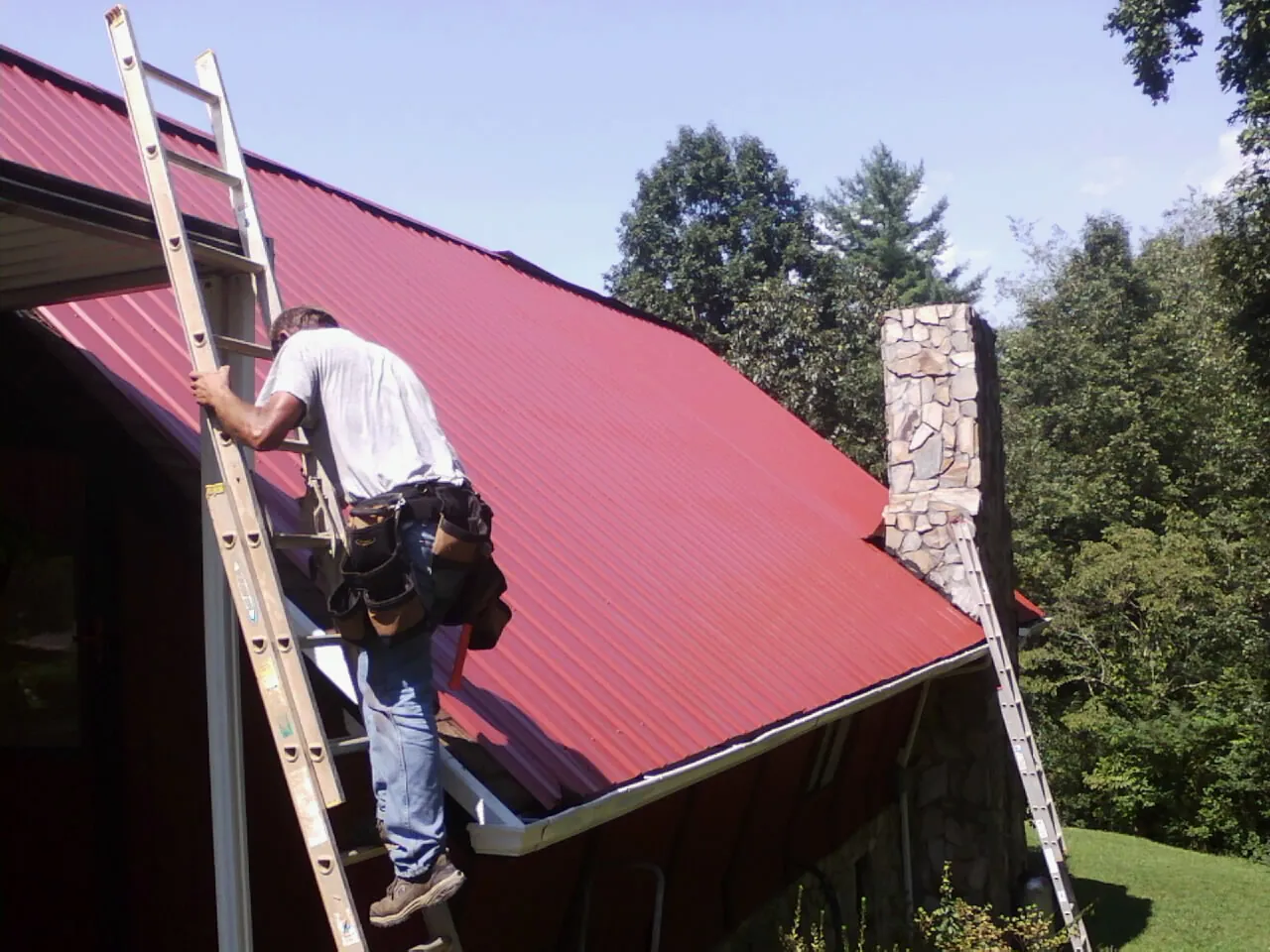 Roofer on a ladder beside a red metal roof and stone chimney — Soffit & Fascia Repair by Roofing Brothers in Tonganoxie